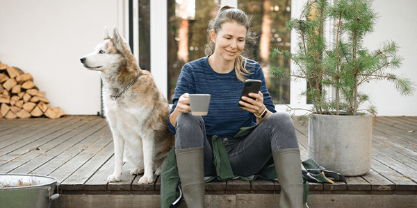 Woman sitting on porch with a dog and a bush to plant, drinking coffee and looking at her phone