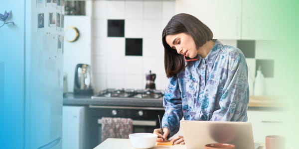 Woman on a phone call holding the phone between her ear and should, while writing something down on a notepad on a kitchen countertop.