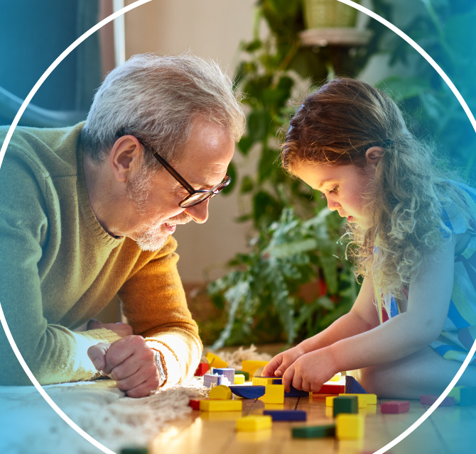 Man and young girl playing inside with toy block on the floor. Indoor plants and furniture in the background.