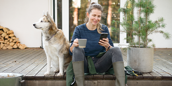 Woman sitting on porch with a dog and a bush to plant, drinking coffee and looking at her phone