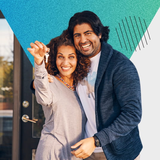 Couple holding each other while in front of home. One member of the couple is dangling keys in front of them.