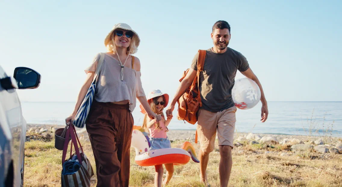 Family at the beach with beach toys. Parents holding toddler by the hands.