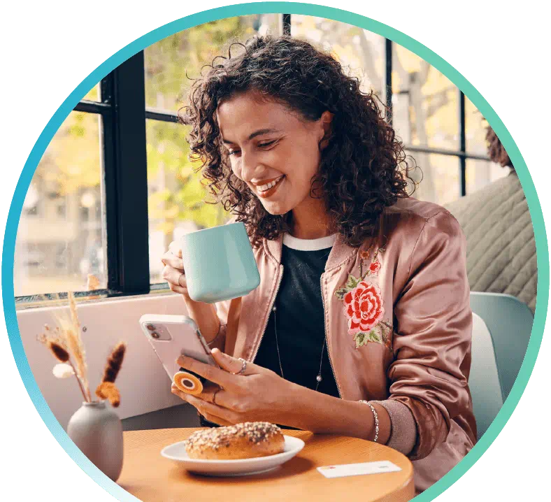 Person drinking out of a mug while browsing there phone and eating a bagel.
