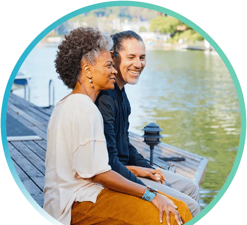 A couple sitting on a deck with their legs dangling off the edge and above a body of water.
