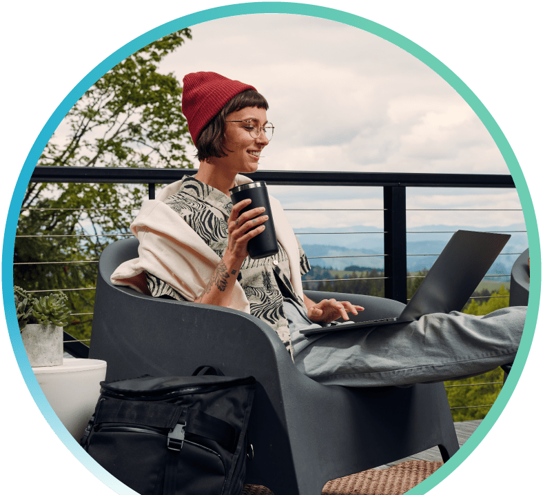 Woman sitting on an outdoor balcony while holding a tumbler in one hand and typing on her laptop with the other hand.