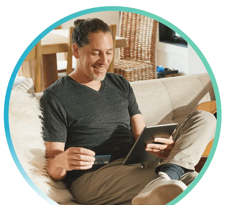 Man sitting on a sofa holding a First Tech debit card in his right hand, and holding a tablet in his left hand.