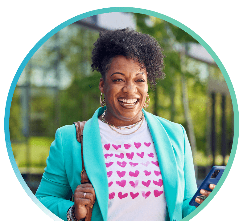 A woman standing outside wearing a teal blazer with a white shirt containing pink hearts on it is standing outside a building while holding her phone.