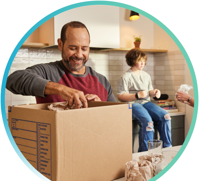 Man unboxing a cardboard box on a kitchen table with a child sitting on the kitchen counter in the background.