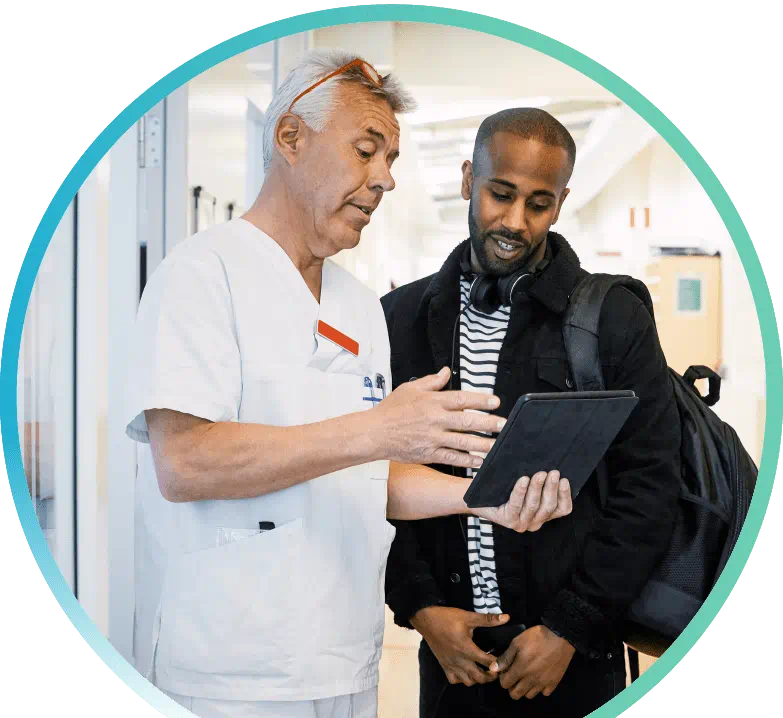 A man dressed in white scrubs is holding a tablet and pointing at it in front of another man  dressed casually with a backpack around his left shoulder.
