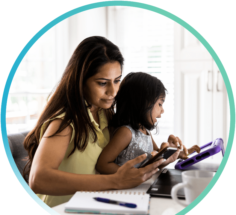 Mother browsing her phone while holding her daughter in her lap who is interacting with a tablet.