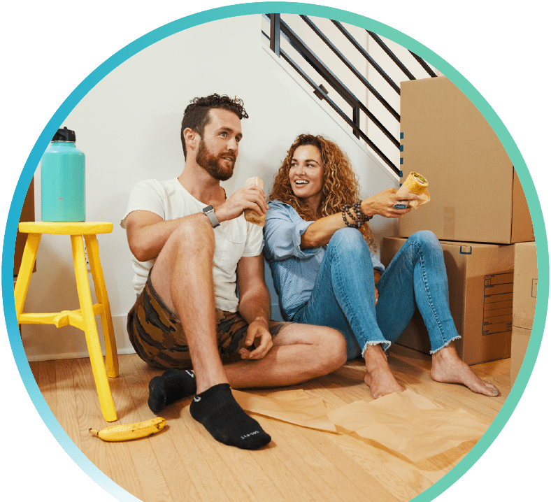 Couple sitting indoors on the floor while eating hoagies with cardboard boxes beside them. 