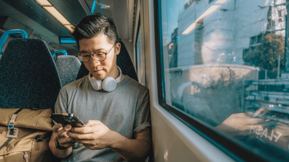 Dark-haired gentleman wearing glasses and headphones around his neck holding mobile phone. Location is on a train and he is reflected in the train window beside him.