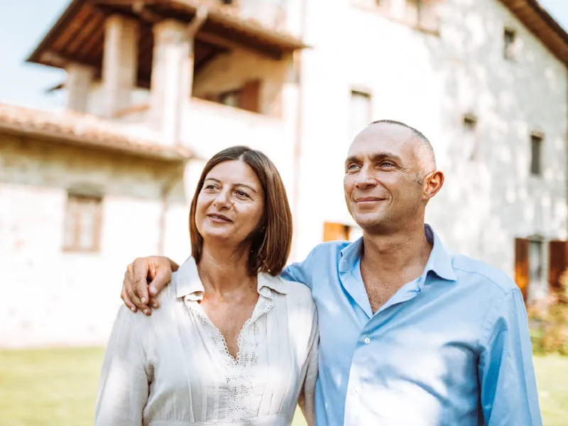 Smiling couple standing outside a house. The woman has brown hair and a while blouse. The man has short grey hair and a blue shirt.