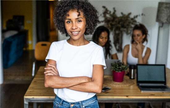 Picture showing three women in what appears to be an office with one of the women standing in the center of the image and in front of the table with her arms folded across her chest. The two other women are in the background, sitting behind the table and looking down at a laptop.