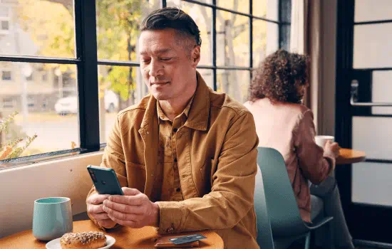 Man sitting in a coffee shop with a mug and pastry on the table in front of him, while he is browsing his phone.