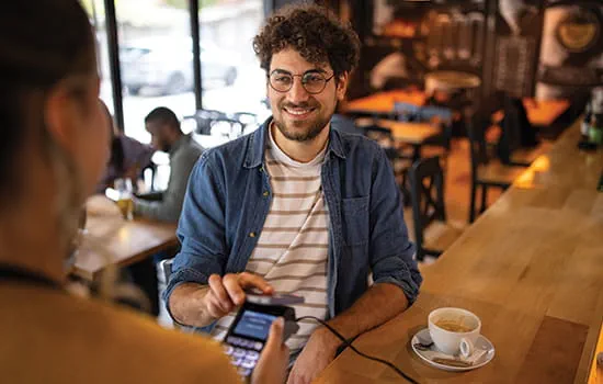 Smiling man with curly brown hair, a beard and glasses touching his phone to a payment scanner at a coffee shop