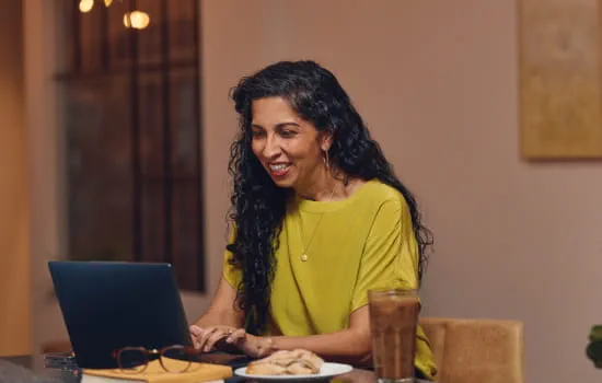 A woman sitting at a table working on a laptop that is open in front of them with a plate of pastries and a beverage on the table as well.