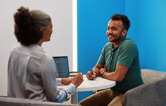 A man is sitting down with a First Tech employee. The man is holding a pen with papers and a laptop on the table as well.