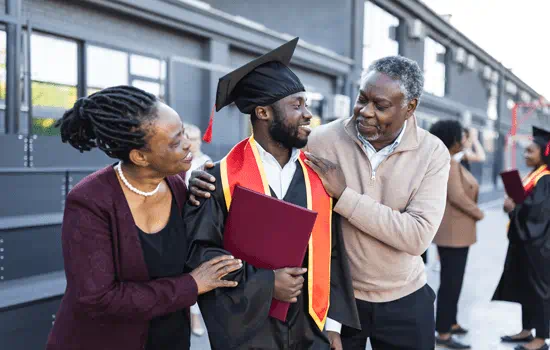 A man wearing a cap and gown and holding his diploma and has his parents beside him.