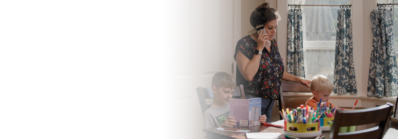 A mother overlooking her children while on the phone. The children are sitting at a table. One child is reading a book, and the other appears to be coloring with markers.