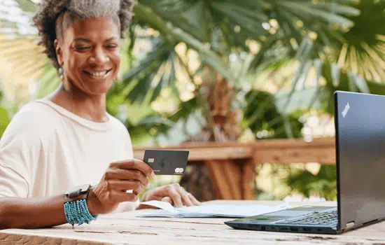 A woman is holding a debit or credit card in her hand while witting outside with their laptop open in front of them.